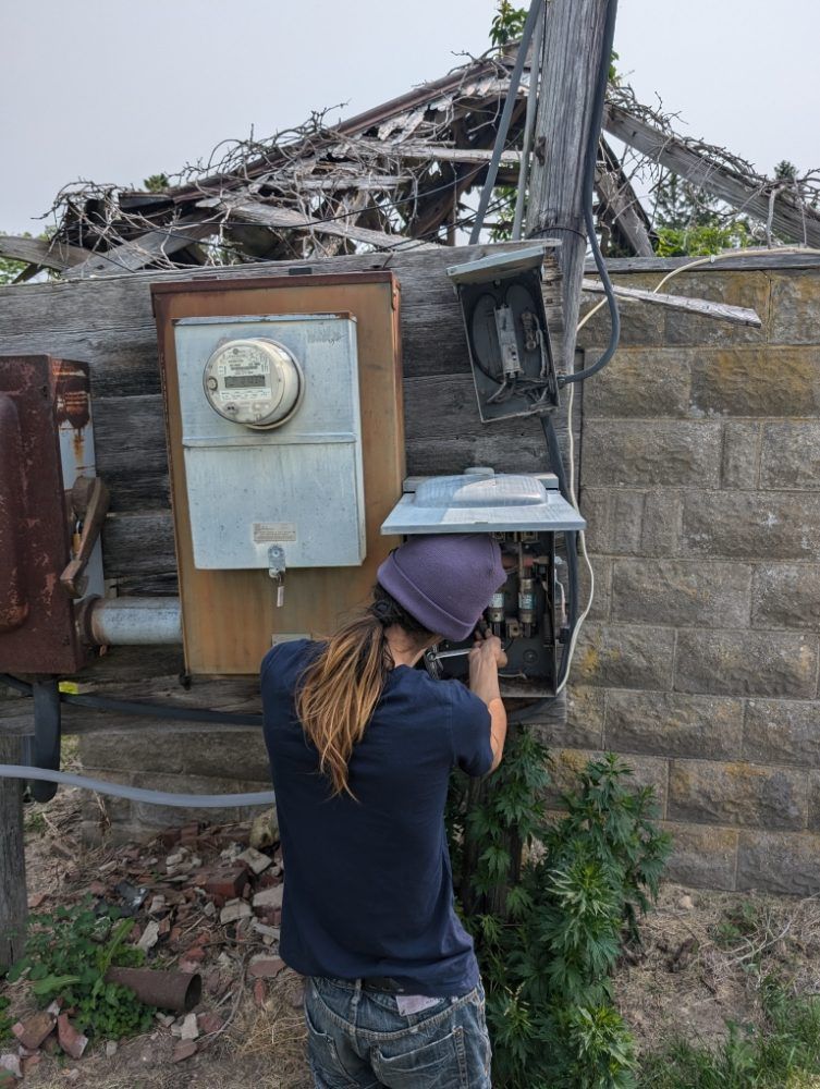 Person with long hair works on electrical box on a weathered stone building. Ruined roof in background.
