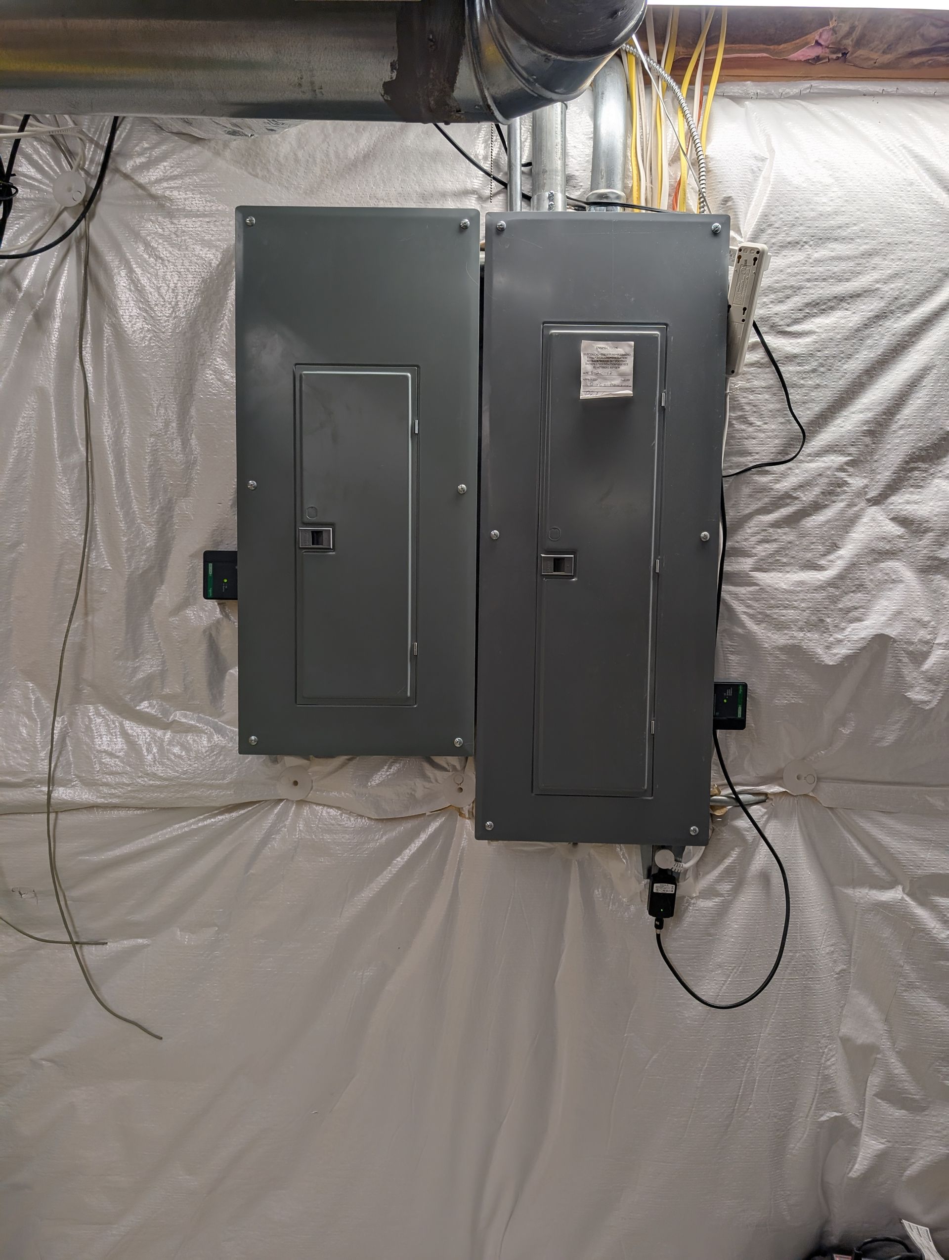 Two gray electrical panels on a wall, with wiring, installed on a white insulated wall.
