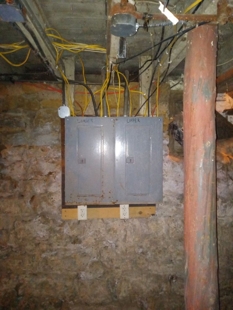 Electrical panels mounted on a wood support in a basement with exposed stone walls and a support beam.