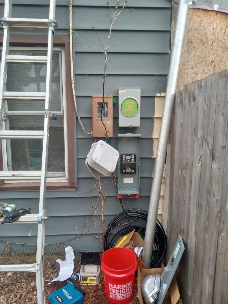 Electrical boxes mounted on gray siding with a ladder, a red bucket, and construction materials.