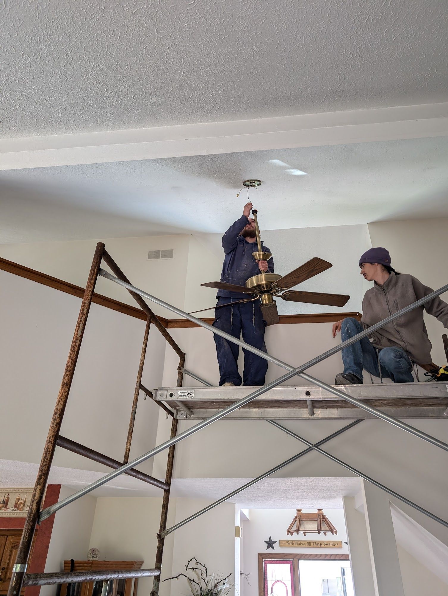 Two men installing a ceiling fan on a scaffold in a white-walled room.