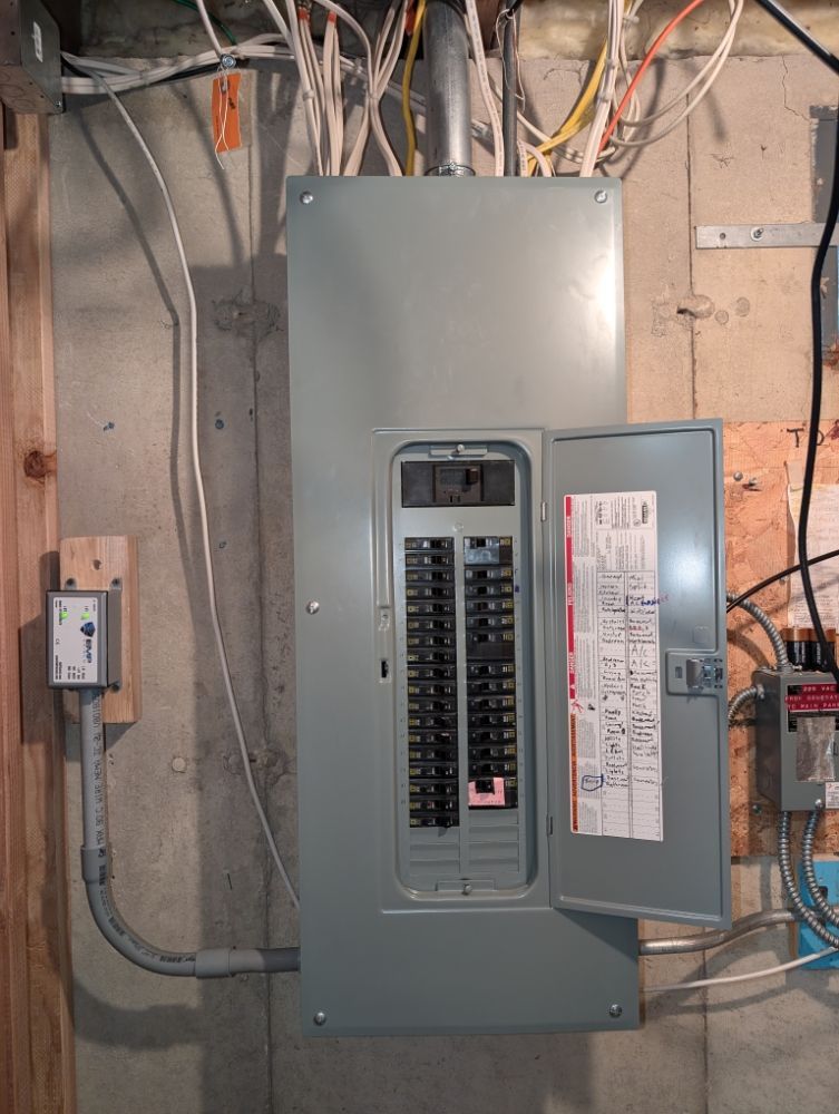 Electrical panel with open door, showing breakers, against a concrete wall with conduits and wires.