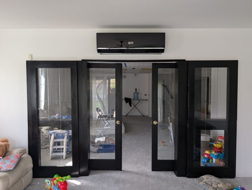 Black framed glass doors on a white wall, with an air conditioner above. A room is seen through the doors.