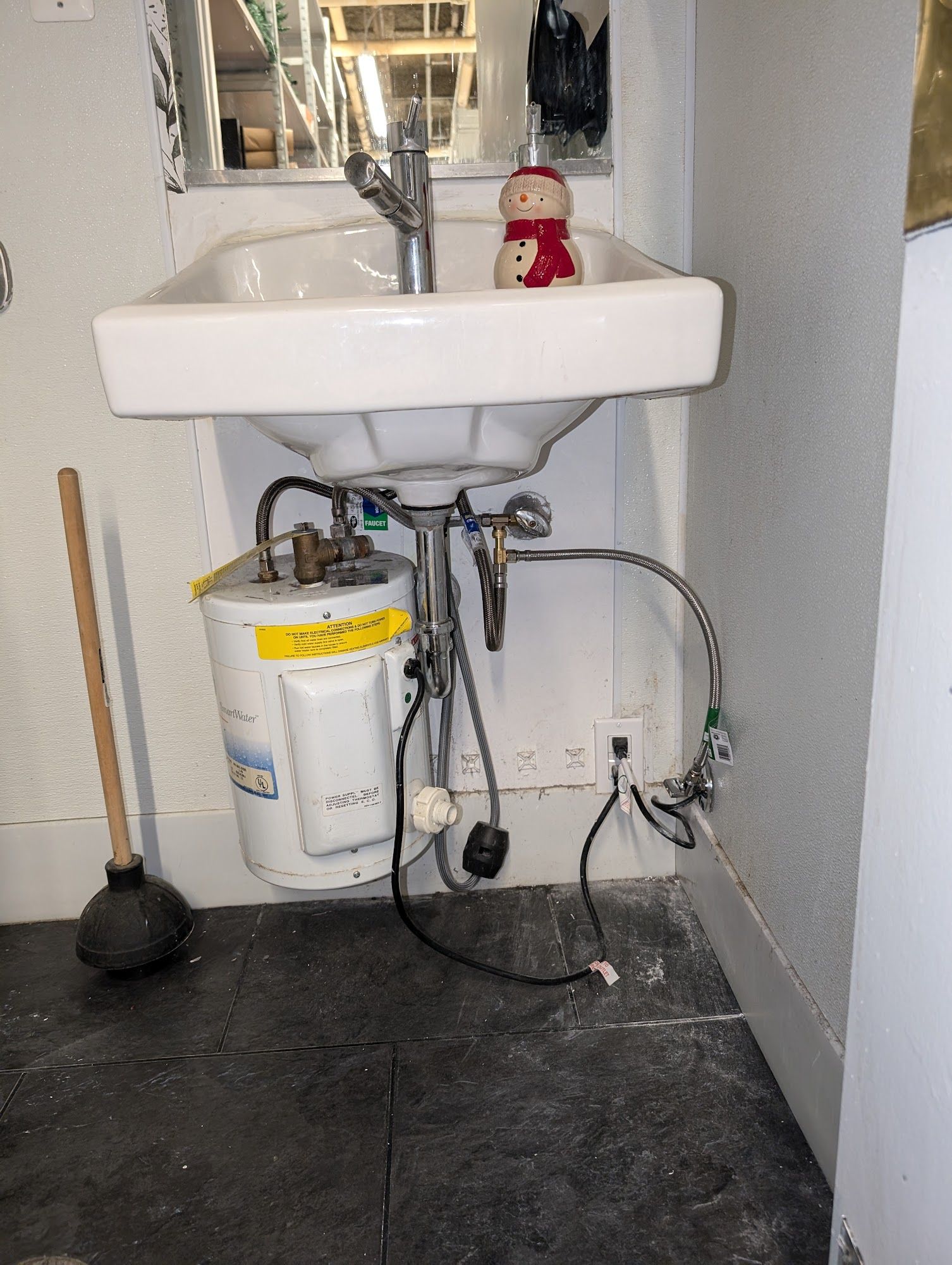 A small bathroom sink with a water heater underneath, plus a plunger and a red snowman.