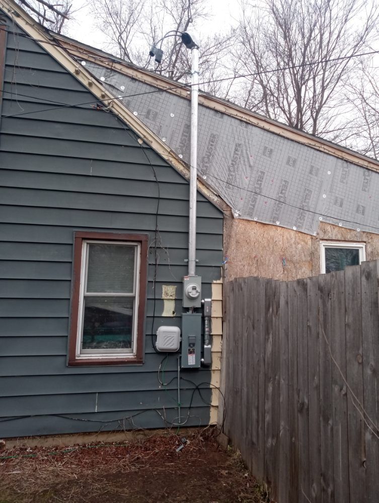 Gray house exterior with a metal chimney, electrical box, and wooden fence.