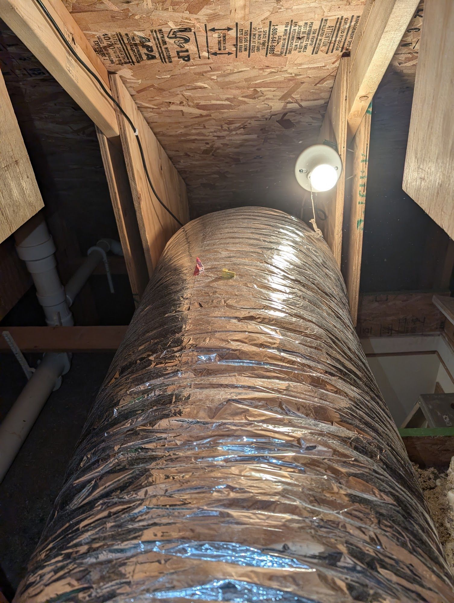 Silver flexible ducting rises vertically through a wooden attic space. A small light bulb illuminates the center.