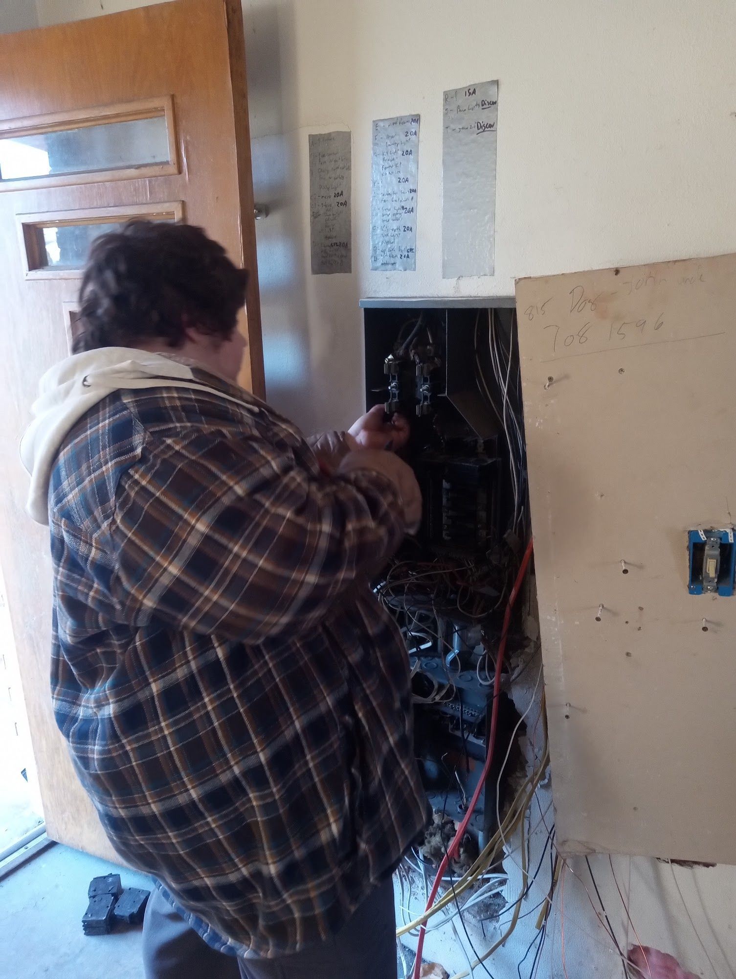 Man in plaid shirt working inside a partially open electrical panel, indoors.
