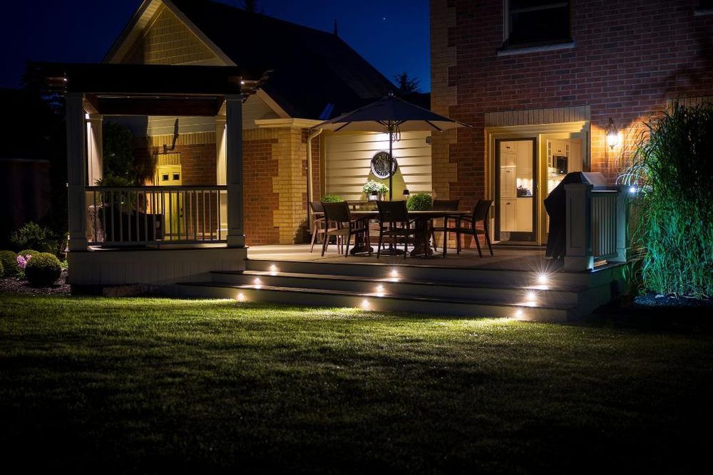 Nighttime backyard scene: patio with table, steps lit by ground lights, next to a brick house.