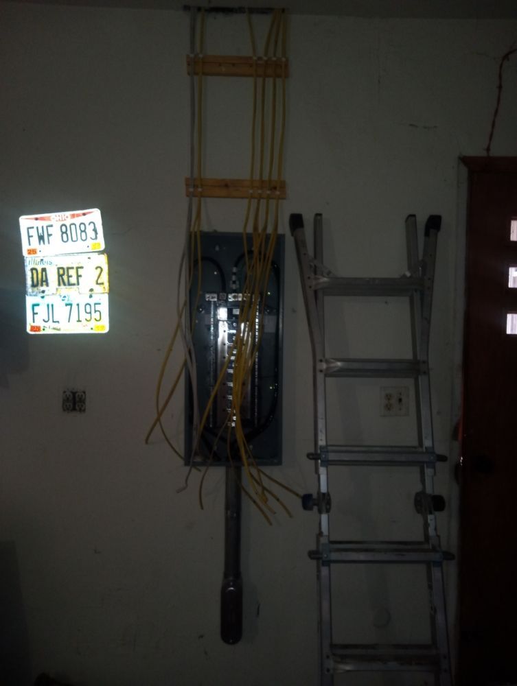 Electrical panel with wiring, ladder, and license plates on a wall.