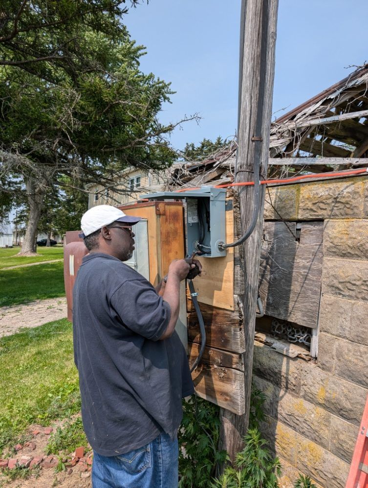 Man in hat working on electrical box attached to a wooden pole next to stone building.
