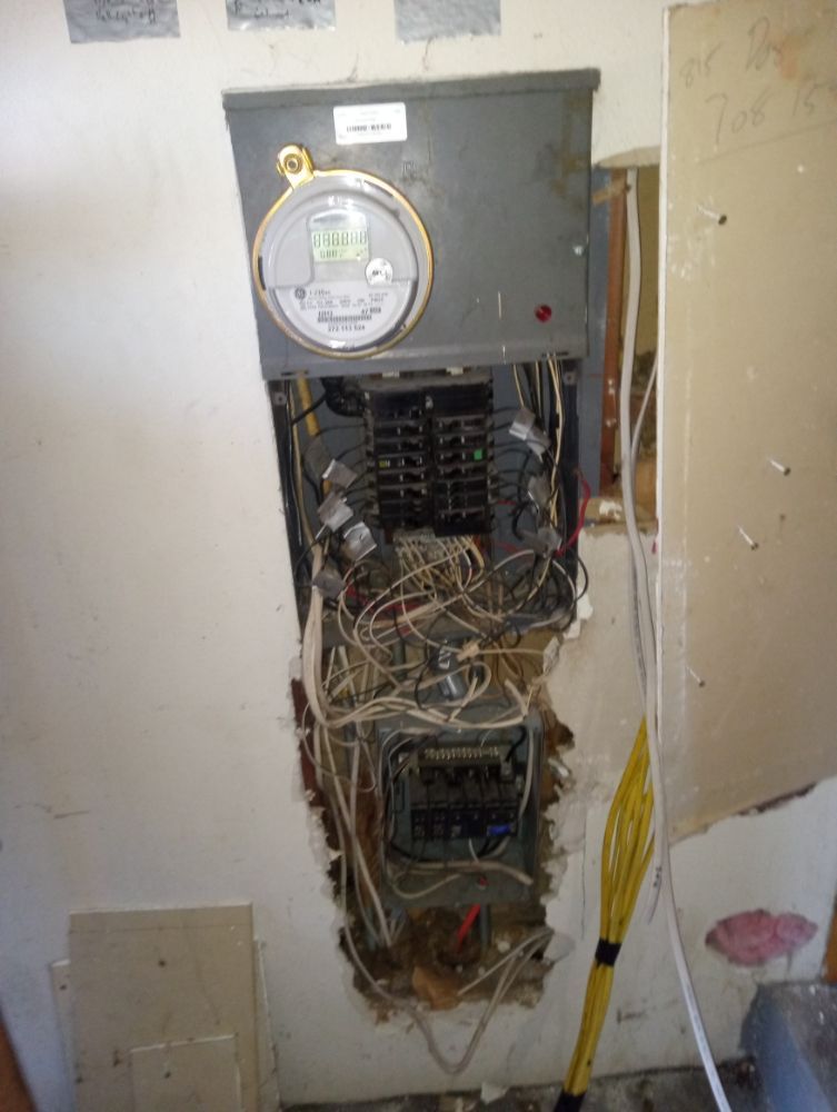 Electrical panel in a wall, wires exposed, meter visible. Damage to drywall around the panel.
