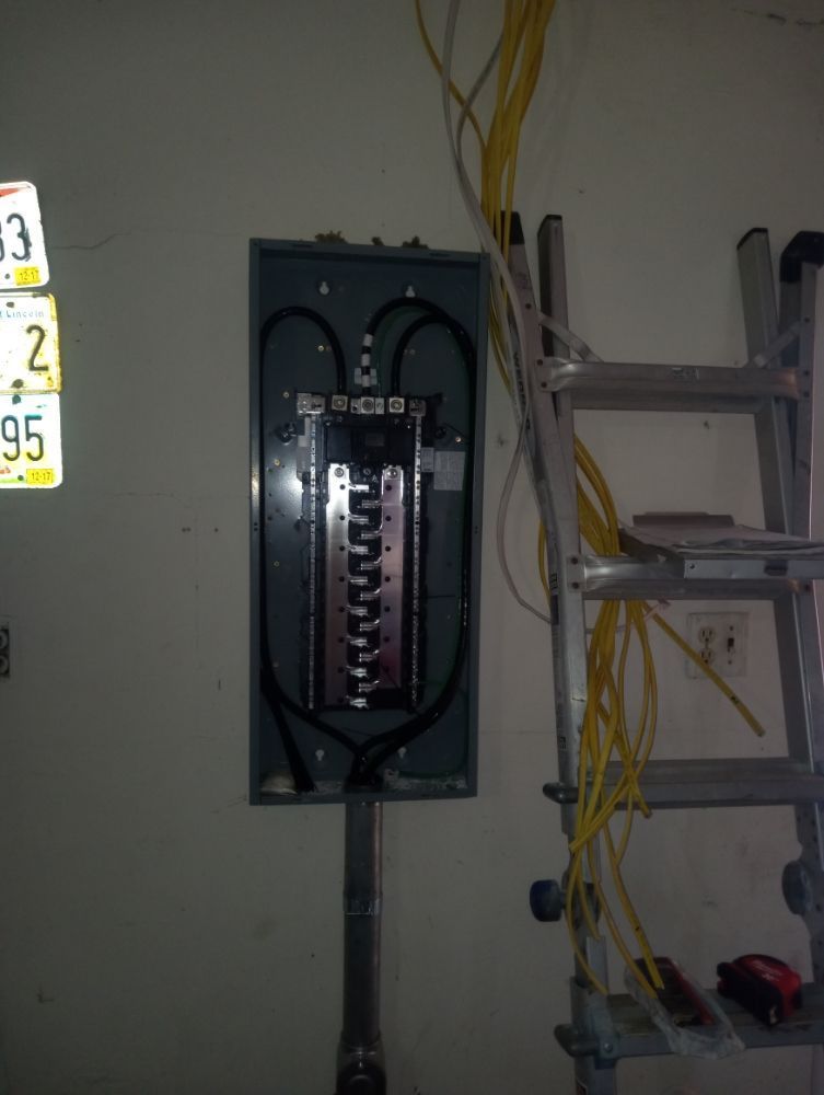 Electrical panel on a wall with wires and a ladder nearby; license plates on the left.
