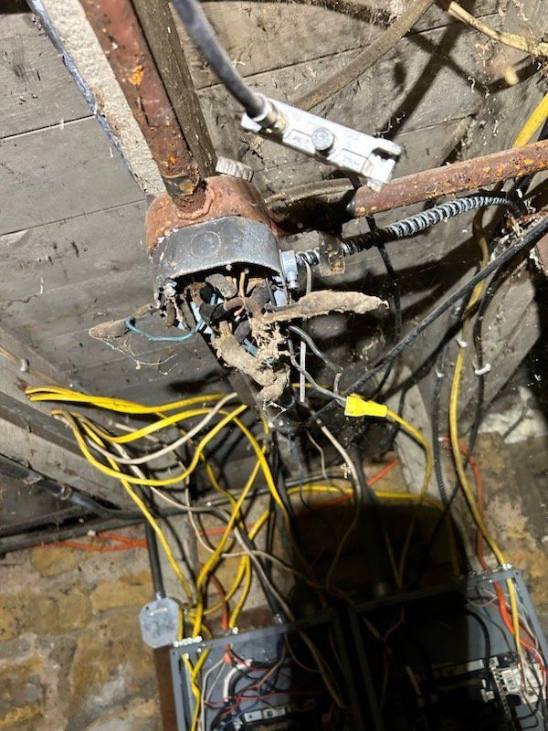 Mess of electrical wiring and components in a dark, old building, with a rusty ceiling fan.