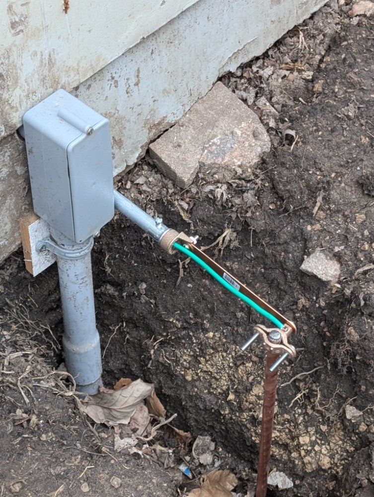 Electrical grounding box and rod next to a building foundation, with green and copper wires.