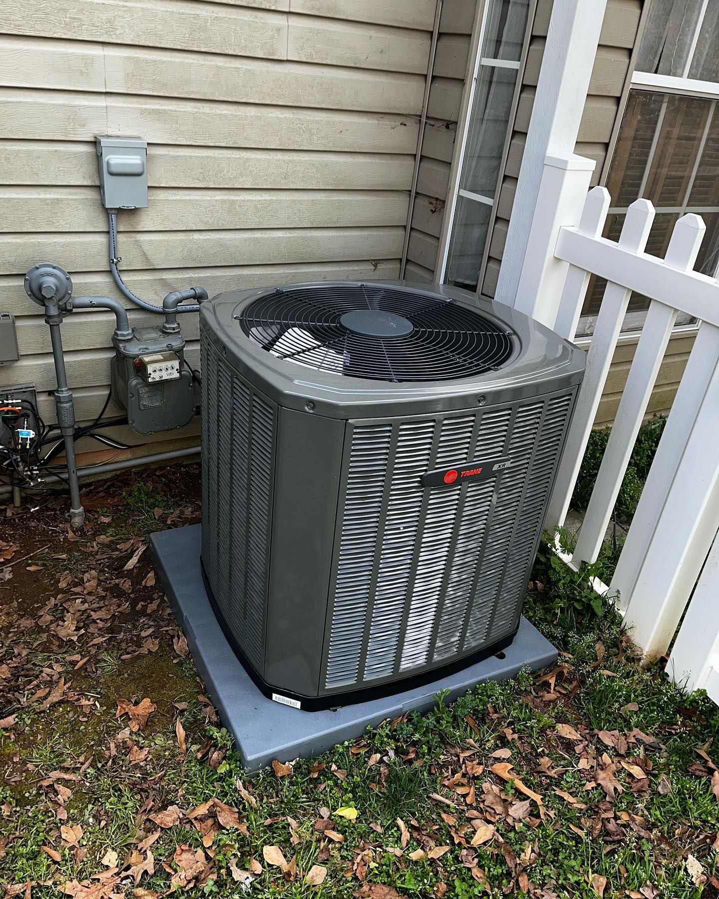 An air conditioner is sitting outside of a house next to a white fence.