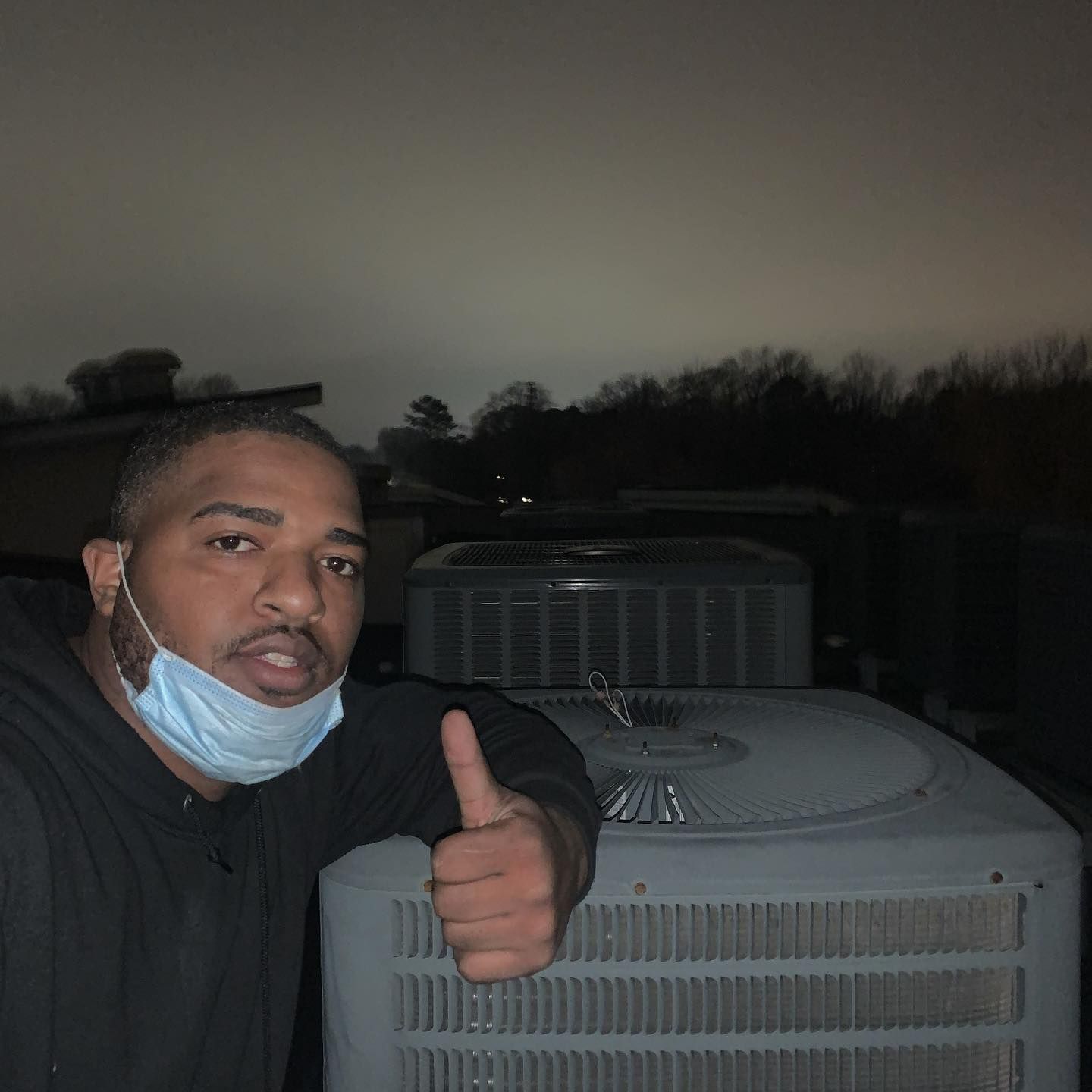 A man wearing a mask is giving a thumbs up in front of an air conditioner.