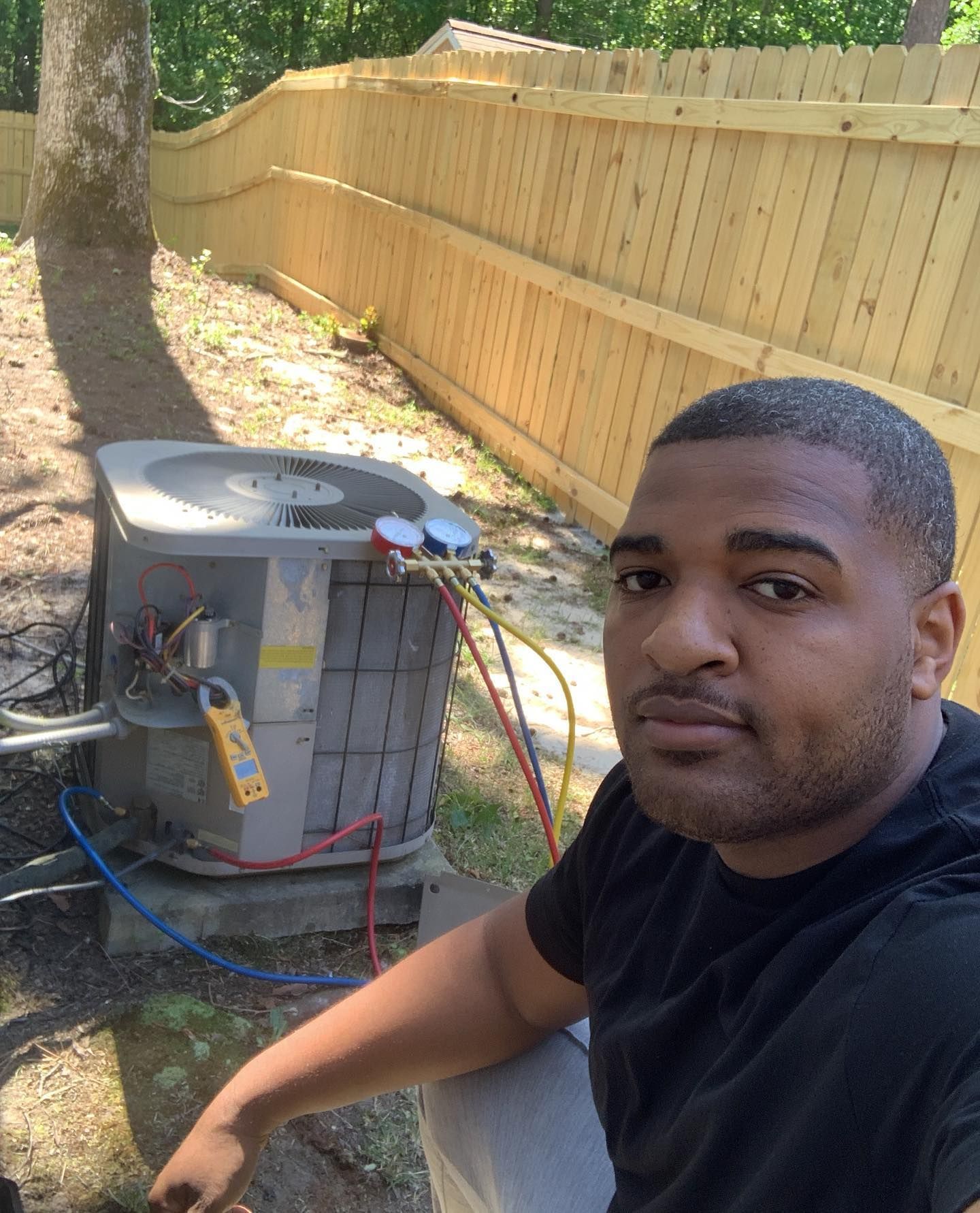 A man is sitting in front of an air conditioner in a backyard.