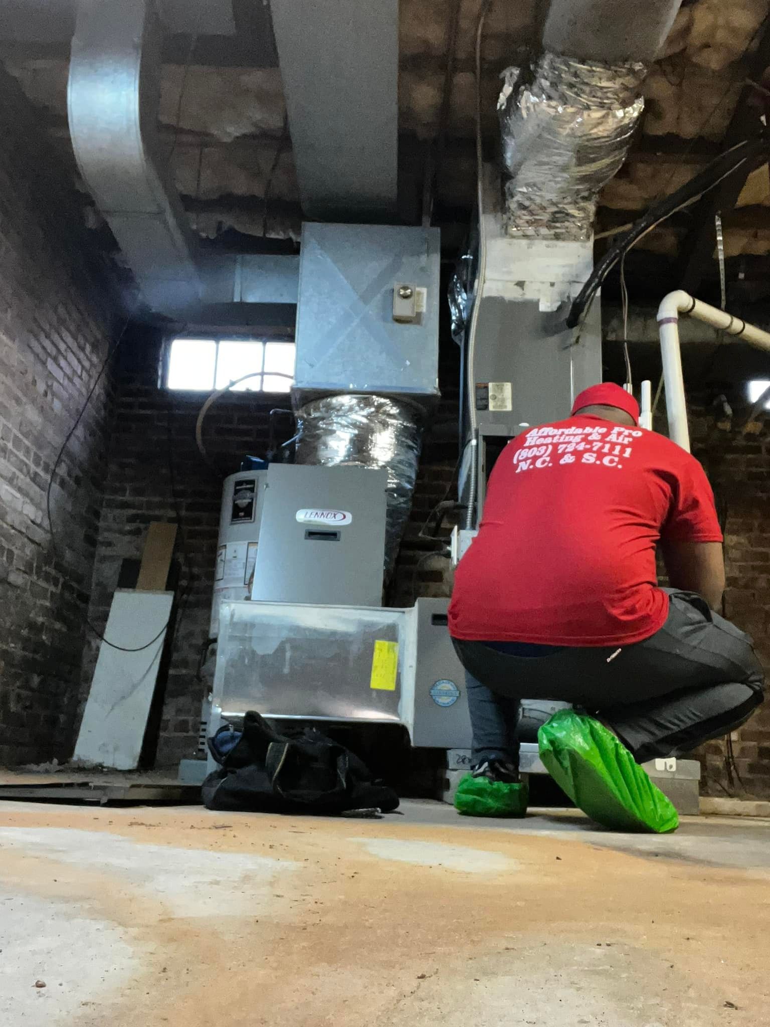 A man in a red shirt is kneeling down in a basement next to an air conditioner.