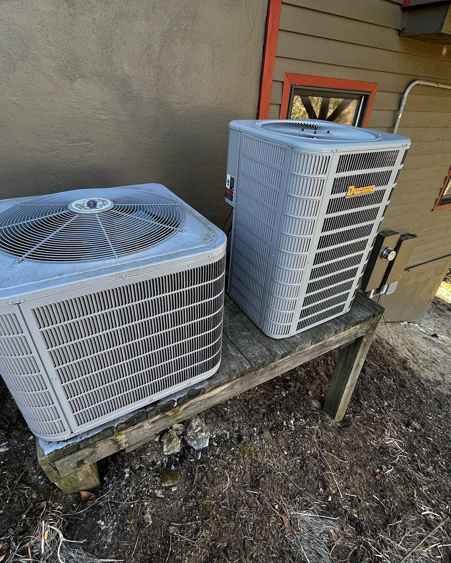 Two air conditioners are sitting on a wooden table outside of a house.