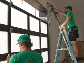 Two workers in green shirts installing a garage door panel, one on a ladder and one below.