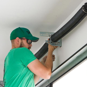 A person in a green shirt and cap adjusting a garage door spring system on a wall.