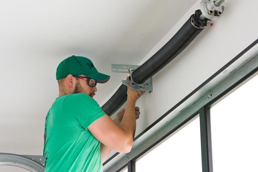 Worker in green shirt and cap installing a black garage door opener rail near the ceiling.