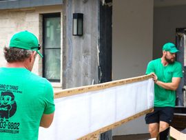 Two workers in green shirts carrying a long white panel outside a building