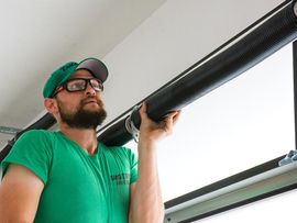 Worker in green shirt and cap inspecting a black window blind or shade overhead