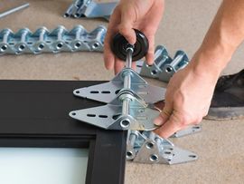 Hands installing a metal clamp on a black window frame with a screwdriver