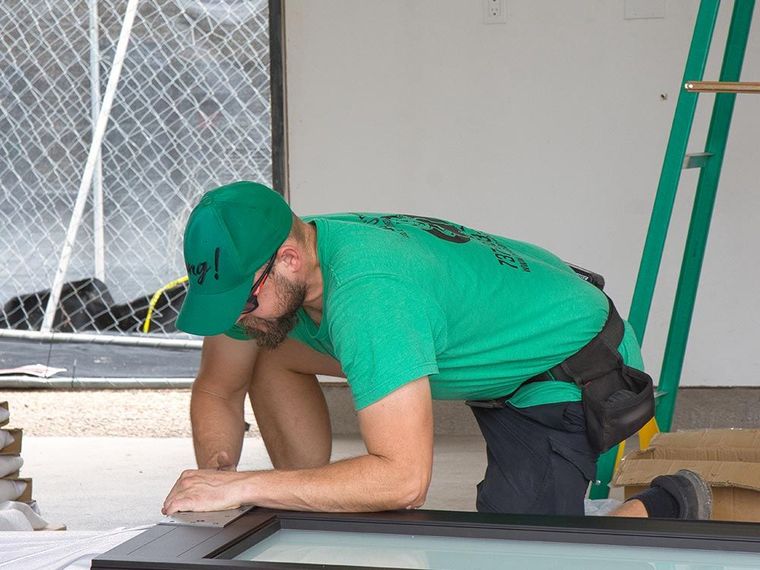 Worker in green shirt and cap kneeling on a floor, using a power tool near a wall and window.
