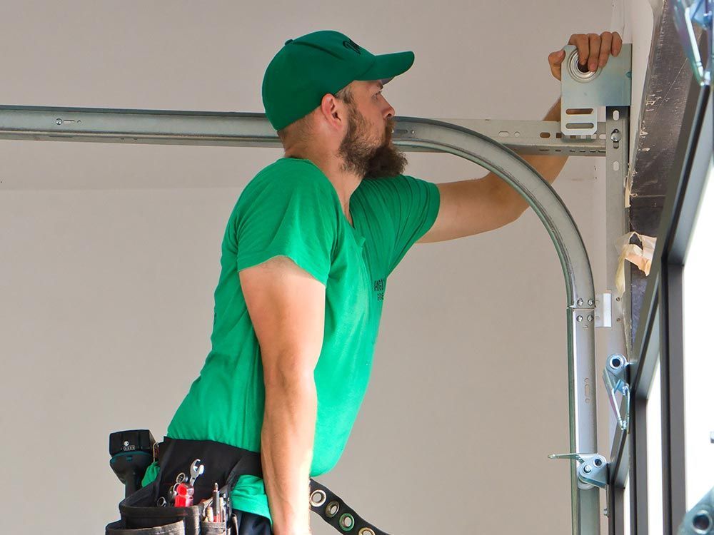 A worker in a green shirt and cap installs a metal garage door track on a wall.