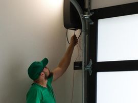 Man in green cap reaching up to adjust a wall-mounted speaker or camera near a window