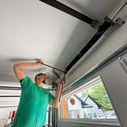 A person in a green shirt works on the mechanical components of a garage door track attached to a ceiling.