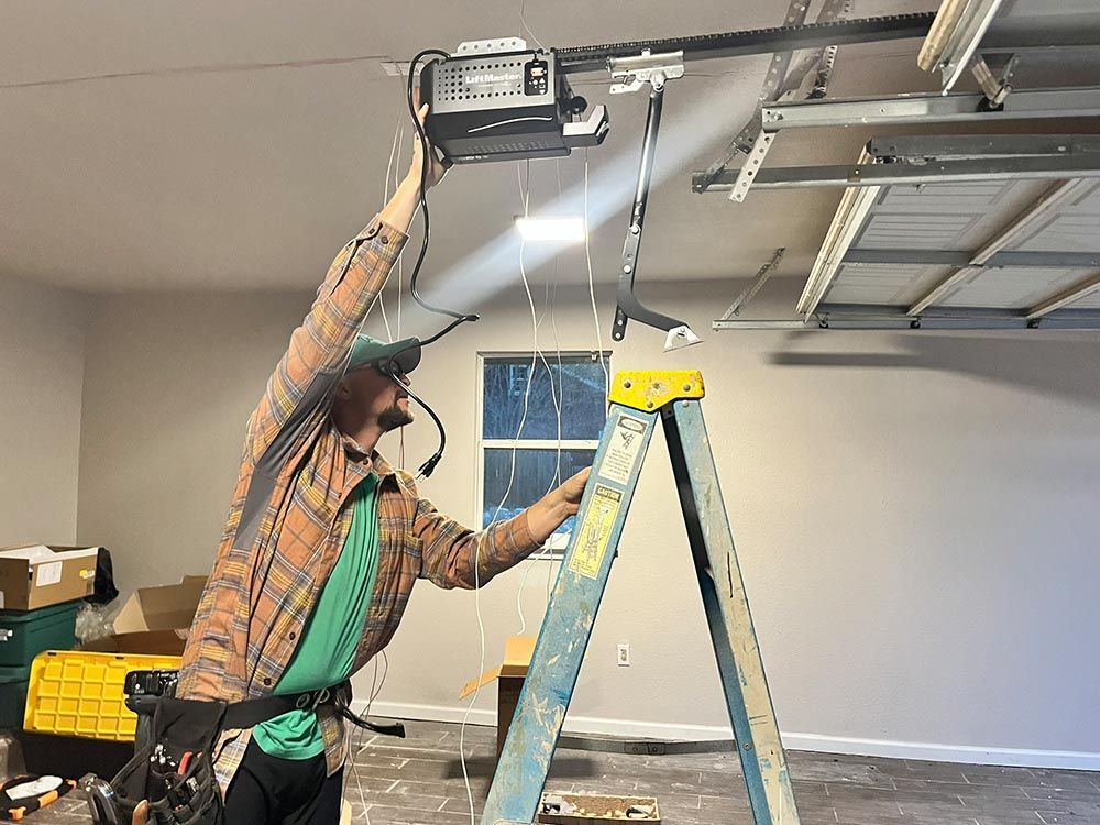 Person in a cap works on a garage door opener while standing on a ladder in a garage.