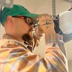 A person in a green cap and plaid shirt working on the wiring of a white garage door opener mounted on a ceiling.