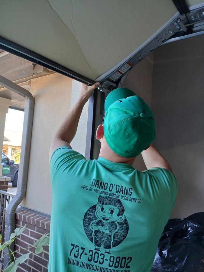 Person in green cap and shirt adjusts a garage door hinge under an awning.
