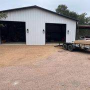 White metal garage with two open bays and a small trailer parked on the gravel driveway.