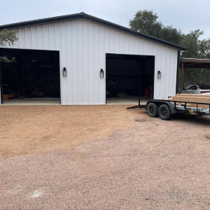 White metal garage with two open bays and a small trailer parked on the gravel driveway.