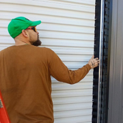 A person in a brown long-sleeved shirt and green cap installs black door-seal weatherstripping on a white corrugated wall.