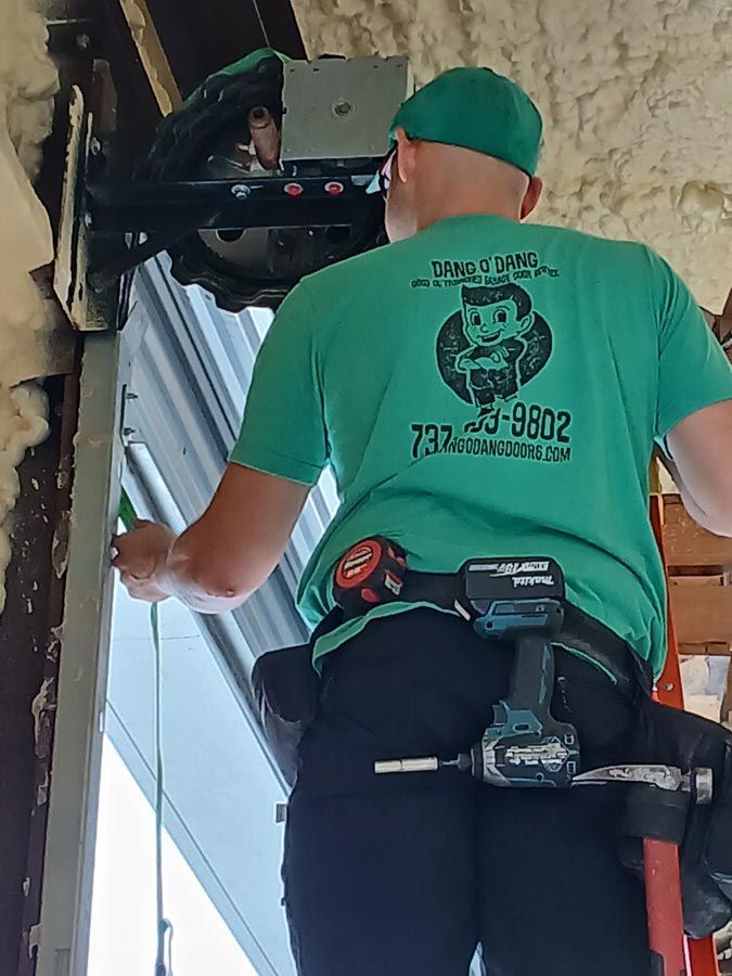 Technician in green shirt and cap works on a window with a power tool from a ladder.