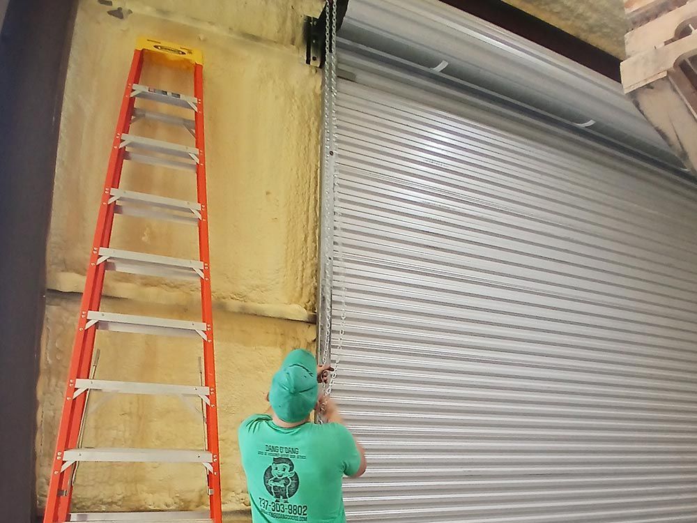 A person in a green shirt works on the side track of a silver roll-up door next to an orange ladder in a storage space.