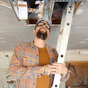 A person with a beard and glasses holds a ladder while standing under an open attic hatch in a workshop setting.