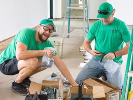 Two men in green shirts and caps examining a small object together against a white background