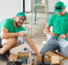 Two men in green shirts and caps examining a small object together against a white background