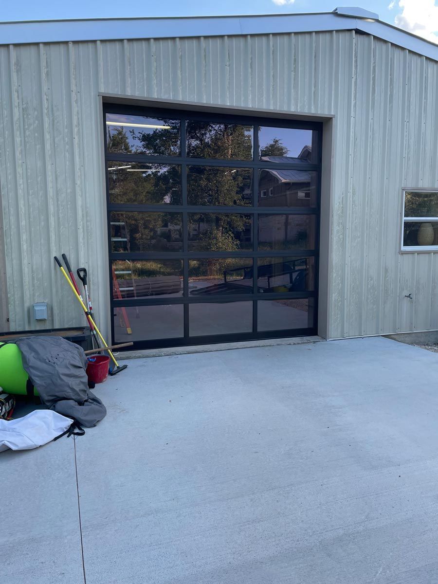 Gray metal building with a large glass garage door and equipment stacked on the left