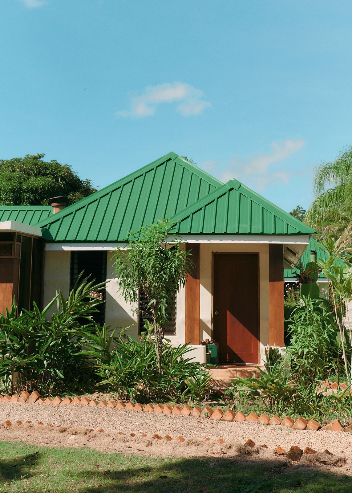Small beige house with a bright green roof, brown door, and surrounding greenery under a blue sky.