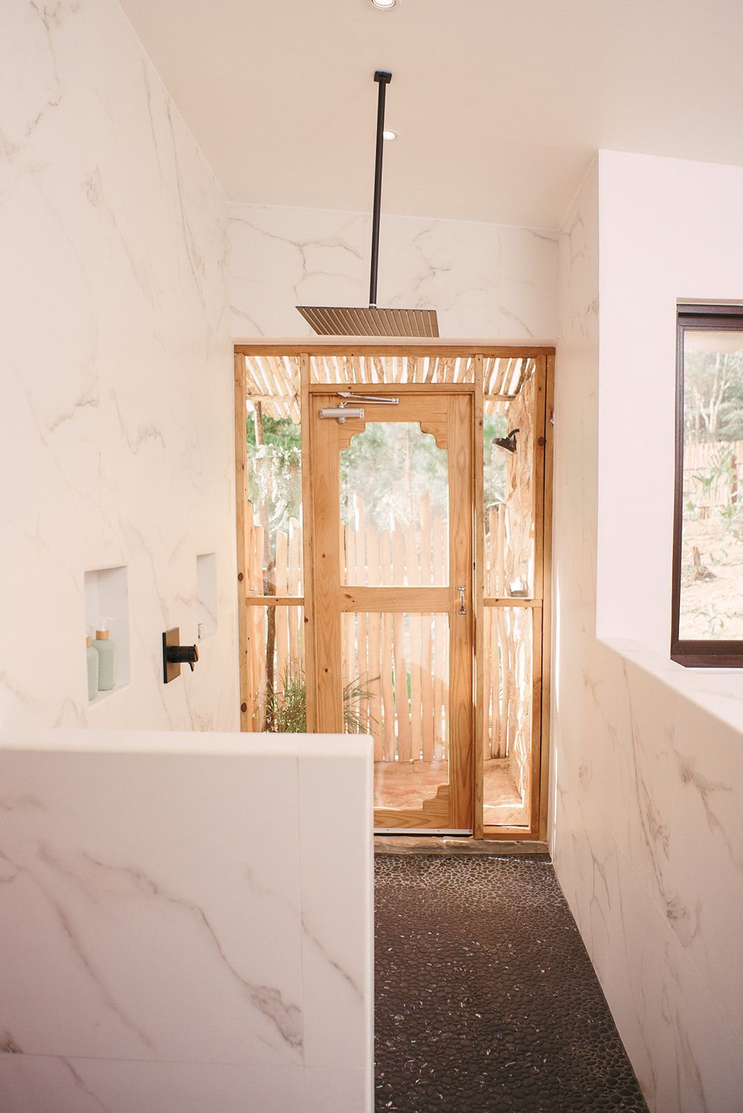 A modern, open-concept shower featuring a rainfall showerhead and a door leading to a private outdoor shower.