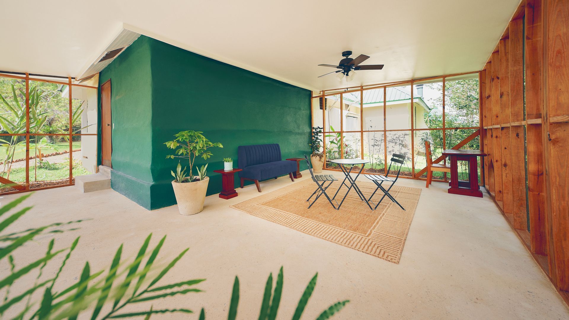 Sunroom with green accent wall, sofa, small table, and plants, with a view of greenery.