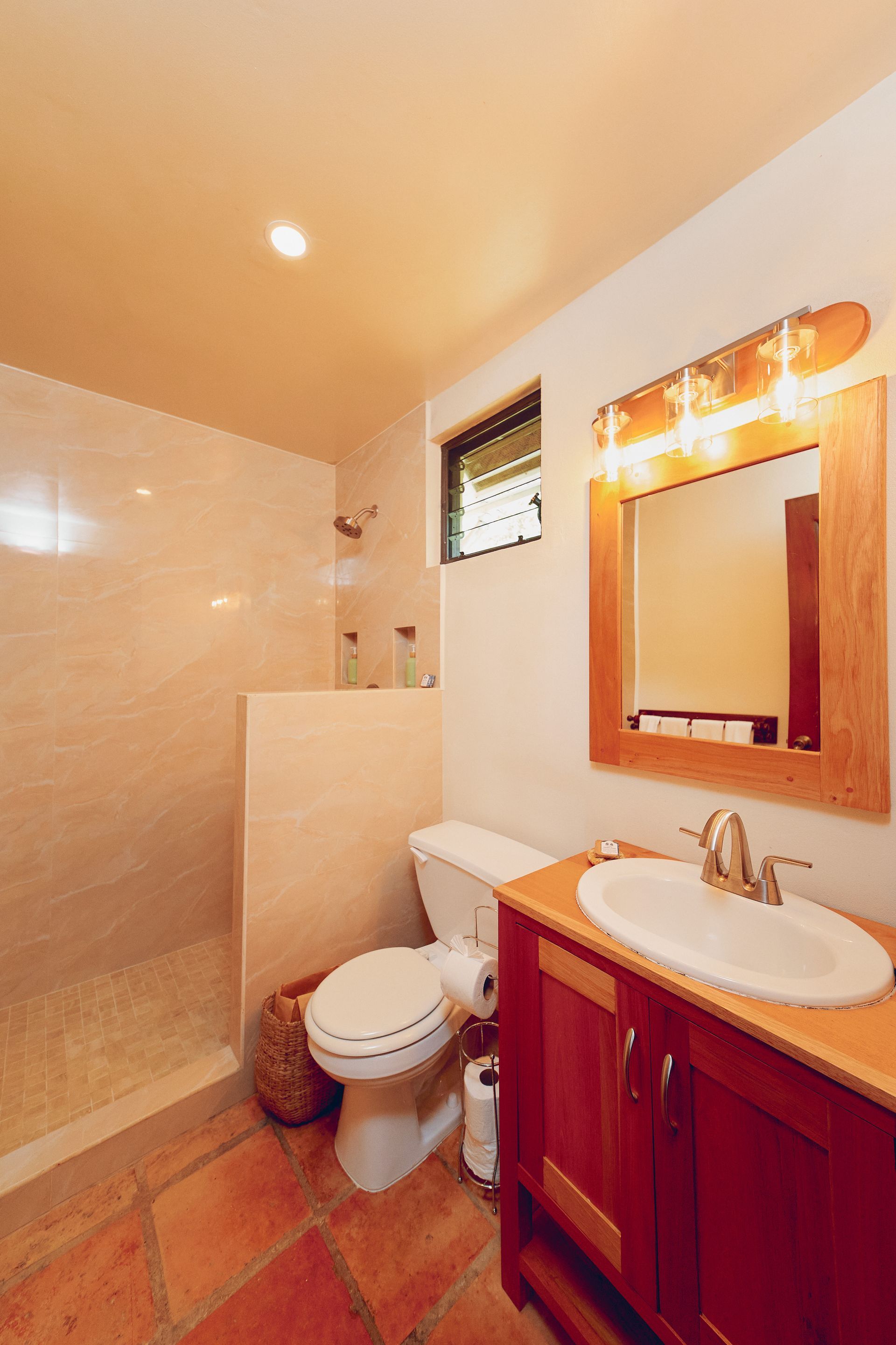 Bathroom with shower, toilet, vanity, and terracotta tile floor; cream-colored walls, wood vanity and mirror frame.