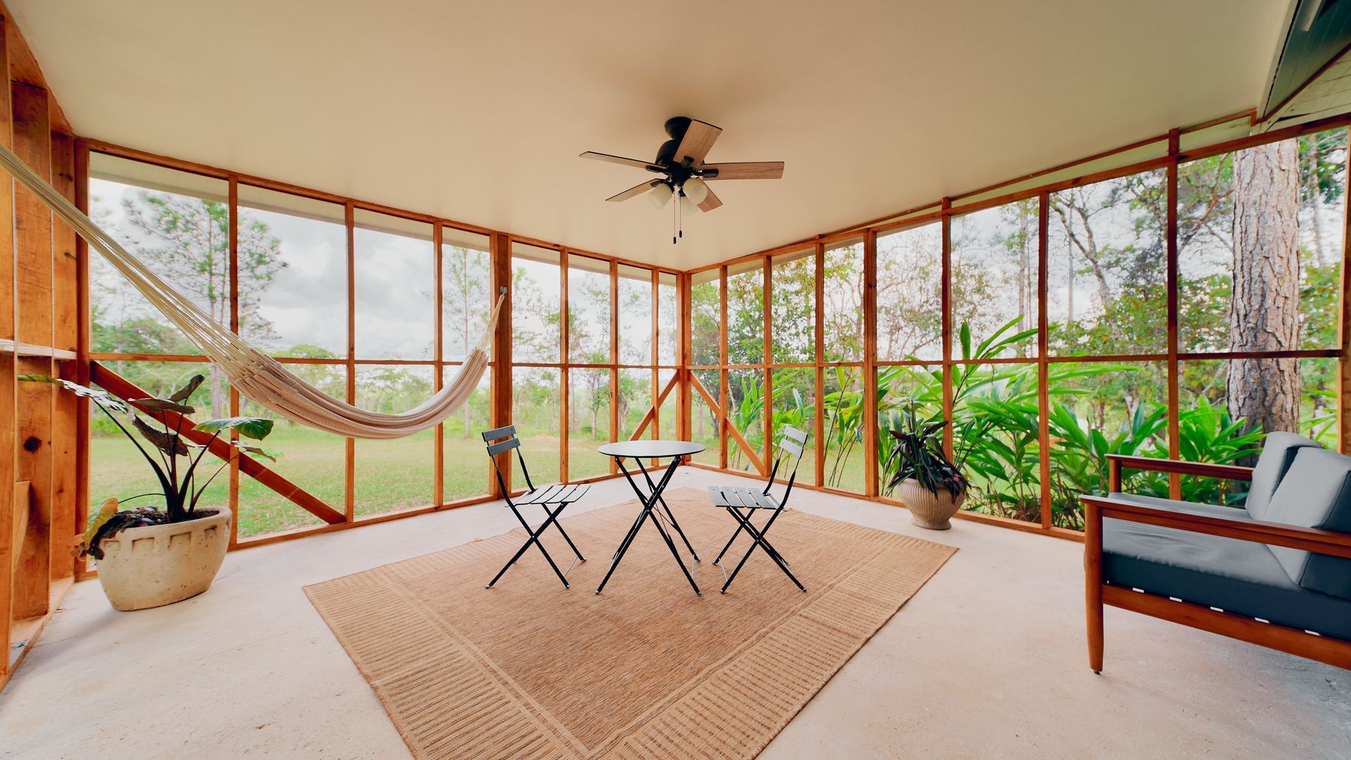 Sunroom with wood frame, glass walls, hammock, small table, chairs, rug, and plants.